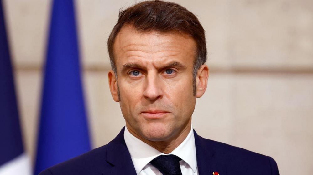 Macron, wearing a blue suit, looks ahead with a stern expression