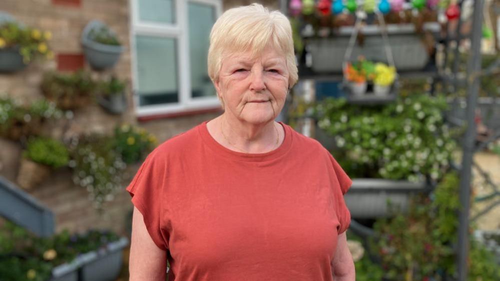 Marilyn Wright, wearing a coral T-shirt, standing in a garden, with flowers and plants behind her. She has short fair hair and is looking straight at the camera.