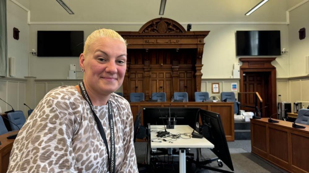Emma Flaxman Taylor has blonde hair tied back in a pony tail. She is wearing a grey leopard print pattern on a white fabric top. She has a lanyard around her neck. She is sitting on a desk inside Great Yarmouth Town Hall council chamber, where there are decorative doors and a wooden canopy feature above the chairperson's bench.