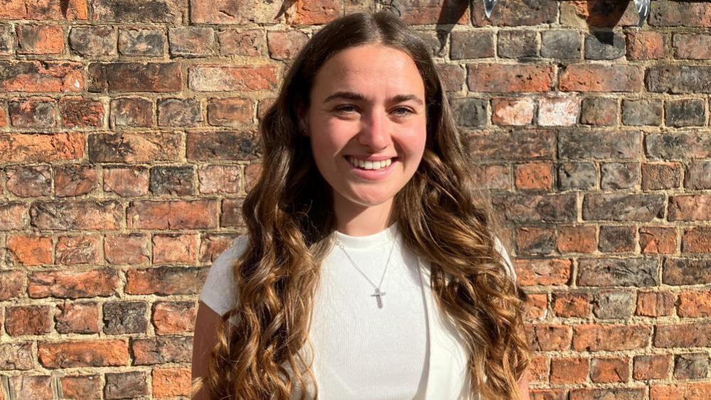 A young woman dressed in white with long, curly brown hair stands in front of a brick wall. She wears a silver necklace with a cross pendant, a white t-shirt and a white waistcoat.