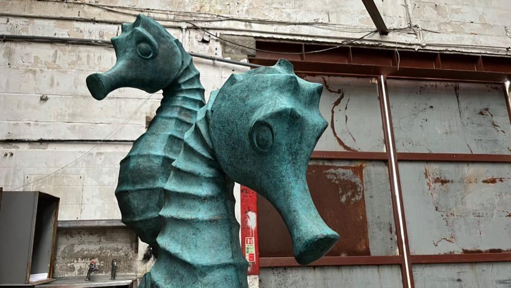 A sculpture of a sea horse, which is a mottled weathered copper colour, standing in a warehouse, with dirty white walls and rusting steel doors behind it.