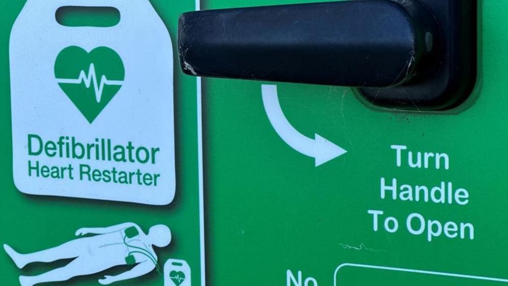 Part of the front of a box containing a public defibrillator. It is green and has a black handle with the instruction Turn Handle to Open. It also features in illustration of a device in use with a Defibrillator Heart Restarter logo above it.