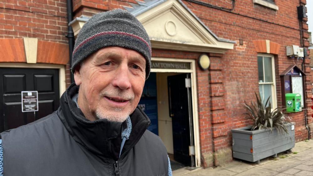 Seamus Elliott is wearing a grey woolly hat, and a black gilet with a blue fleece beneath. He has a goatee beard, and is standing outside the entrance to Aylsham Town Hall.