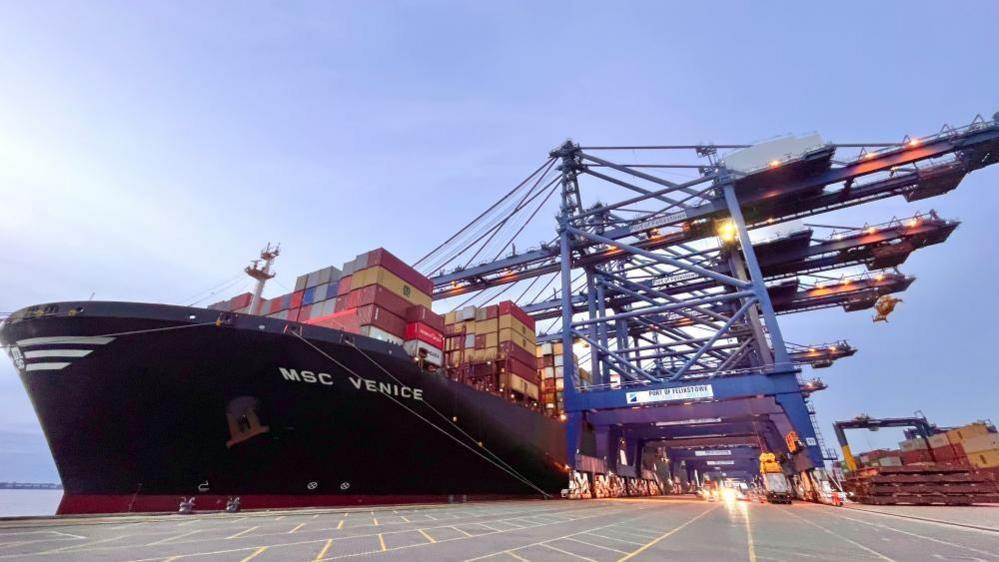 A picture of a large container ship docked at the port of Felixstowe. The ship has a number of cranes hanging over, hauling containers on to it. The ship is MSC Venice. 