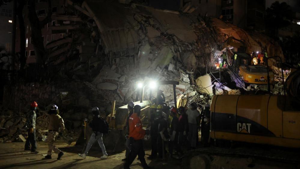 A team of emergency workers using heavy machinery to search a collapsed building, lit by bright headlights in darkness