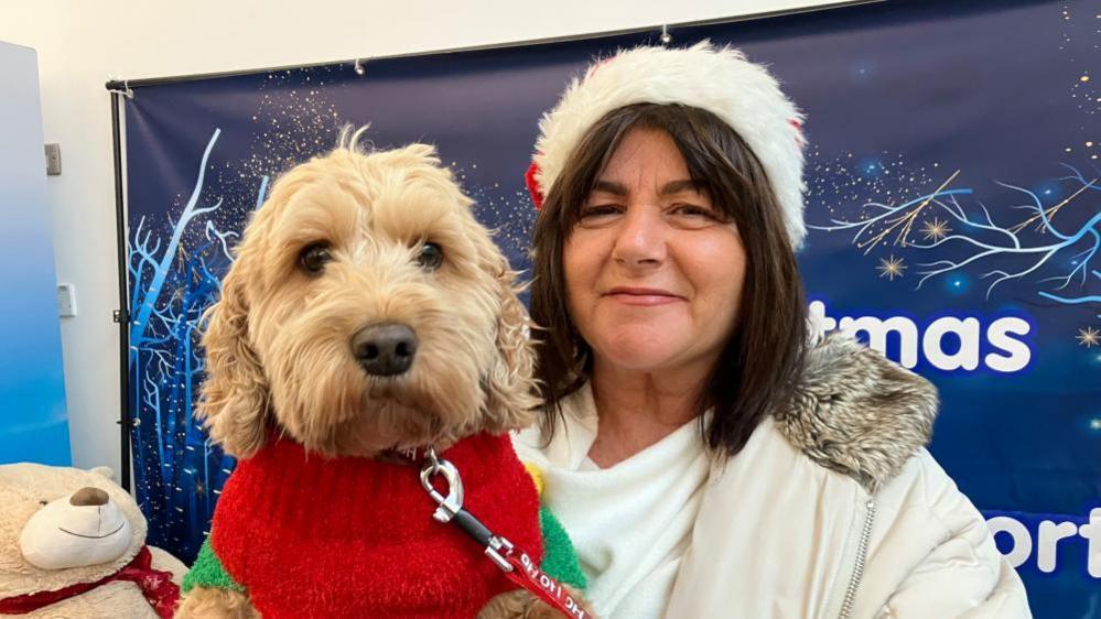 Deirdre has short brown hair and is wearing a Santa hat. She has a beige coat on. She is holding her dog Skye is blonde and wearing a red Christmas jumper.