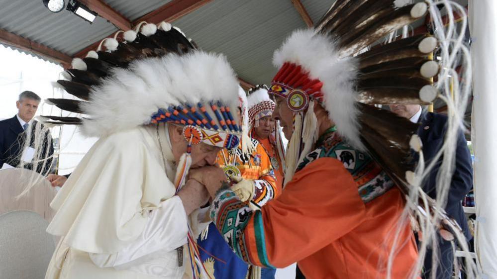 Pope Francis wearing Native regalia and kissing the hand of a chief