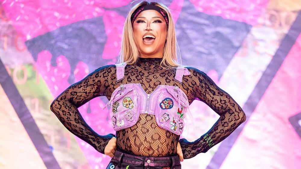 Drag queen Saki Yew is on-stage at Manchester Pride. She is wearing a lace top with long sleeves and a small pink waistcoat over the top of it, covered in brightly-coloured badges. She is open-mouthed, looking out from the stage. She is wearing a black headband with pink letters on it and has long, blonde hair.