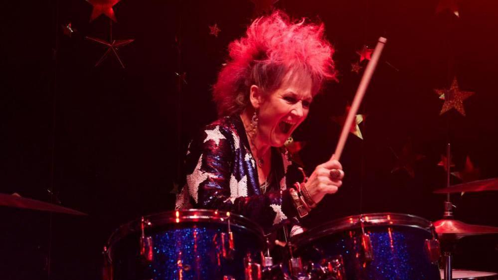 Lorraine Ashbourne as Jess in still from the drama Riot Women. A middle aged woman plays drums in a punk band with spikey hair and a sequin top.