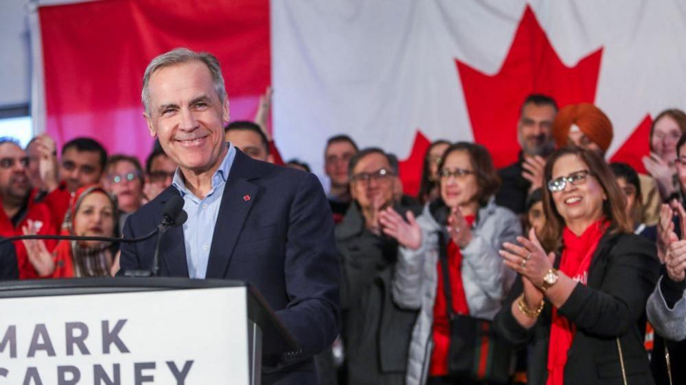 Former Bank of Canada and Bank of England governor Mark Carney stands before a podium with a crowd and a Canadian flag in the background. He looks to the right and is smiling. 