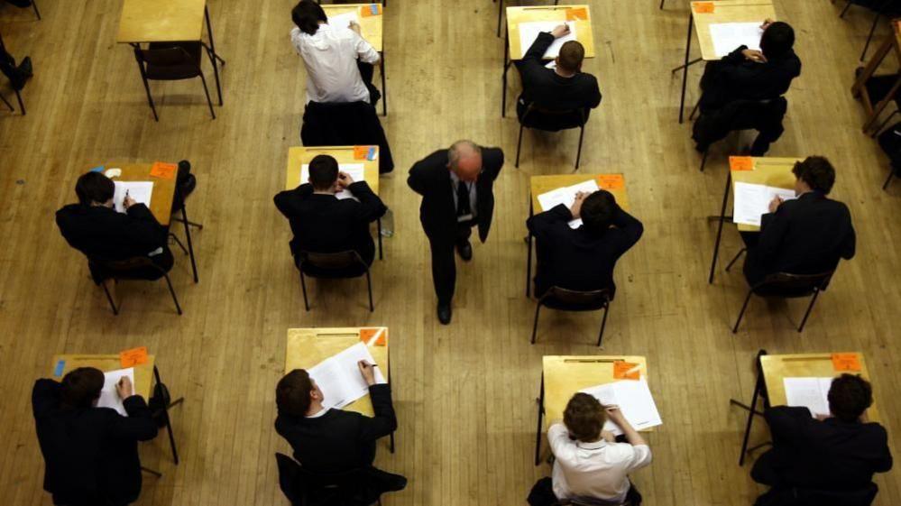 Aerial shot of 11 male pupils sitting at desks in a hall during an exam. They are all wearing black blazers, with the exception of two who have removed their blazers to reveal white shirts. A bald male invigilator, in a dark suit, is walking down the centre of the aisle and looking down to his left.   