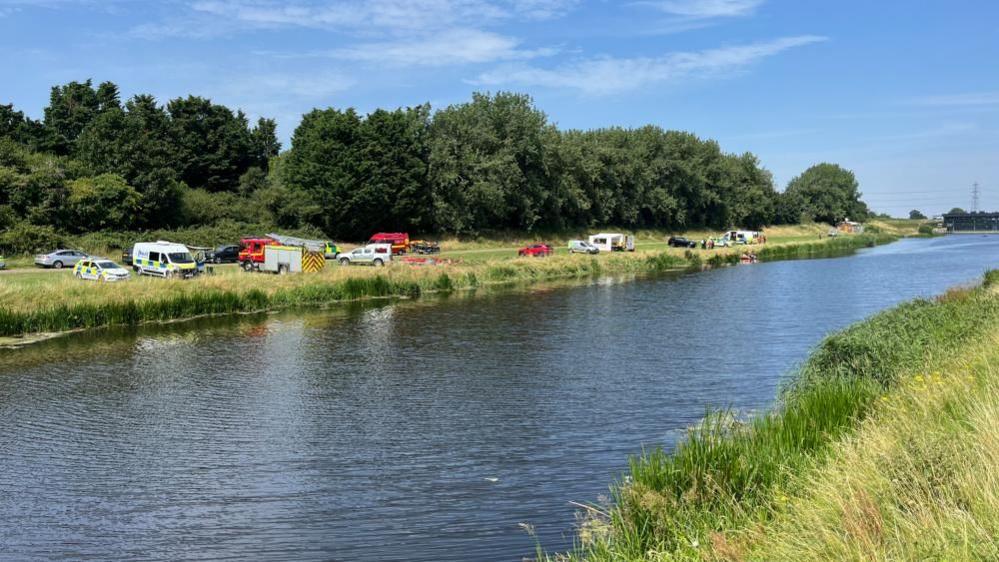 Ambulance, police, fire and rescue appliances and cars and a caravan parked on the far bank of the river, with green banks either side and a row of trees behind on the far side. At the extreme right of the image is a sluice gate structure.