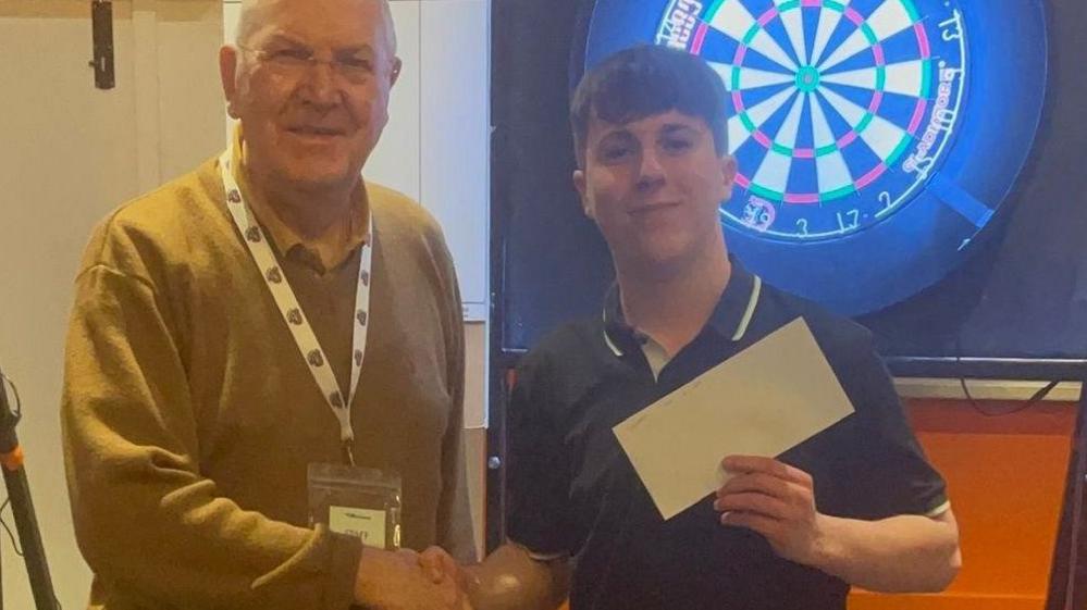 A man is shaking hands with a boy, who is holding a cheque. Behind them is a darts board.