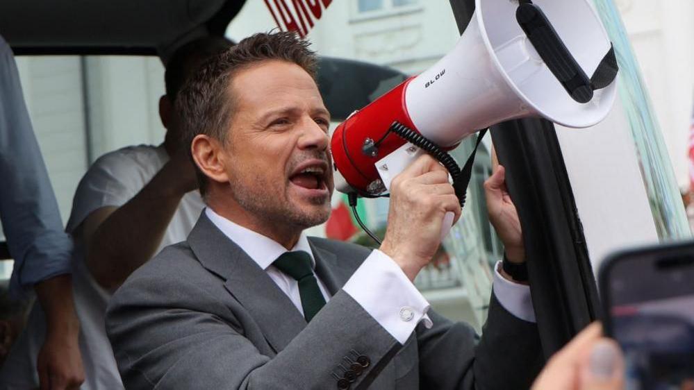 Rafal Trzaskowski wears a suit, stands in a bus doorway, holds up a megaphone and speaks into it