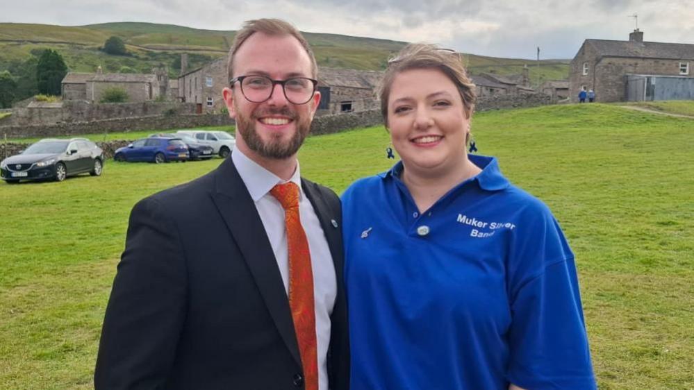 A 30-year-old man wearing a suit and orange tie, next to his 27-year-old sister who is wearing a blue polo shirt reading "Muker Silver Band".