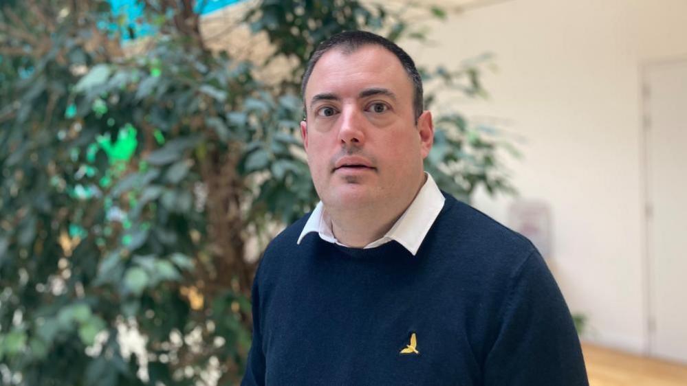 Mike Hallam with short dark hair staring at the camera. He is wearing a blue sweater with a bird logo and a white shirt. He is in front of a large plant and a white wall.