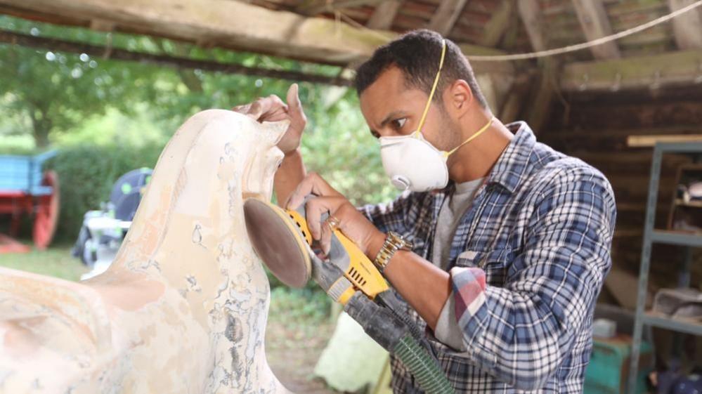 Will Kirk wearing a mask and working on a white rocking horse with a circular saw