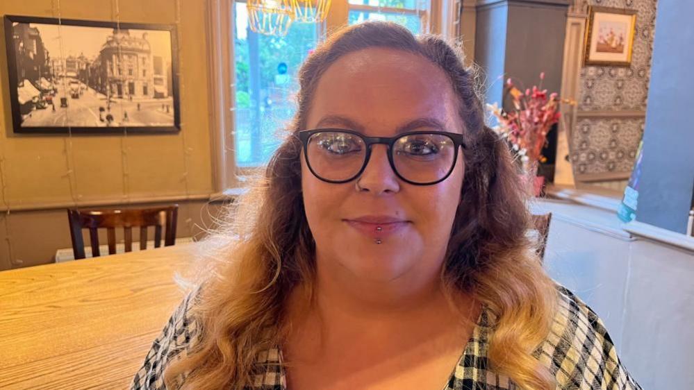 Katie with long fair hair and dark brown glasses, wearing a blue and white checked top in a house.