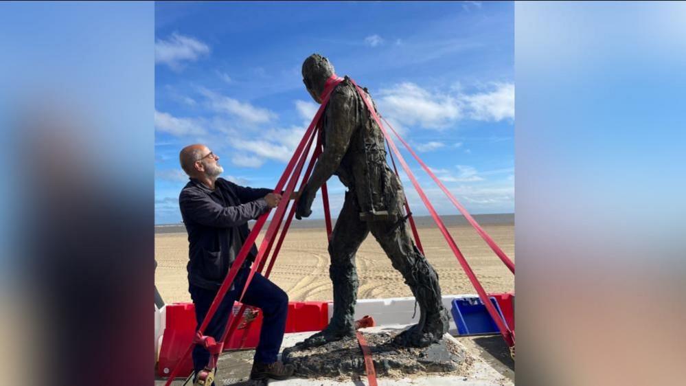 Laurence Edwards with grey hair and a beard puts a bronze statue of a walking man in place on a platform on a beach. There are red straps securing the statue to the board.