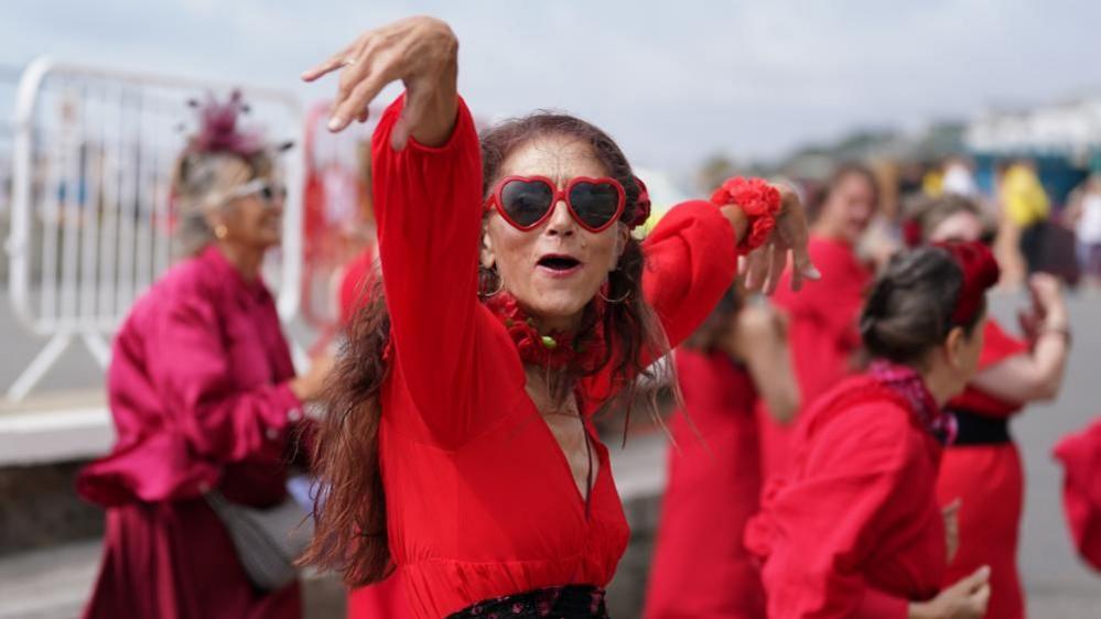 A woman dressed in a red dress with heart-shaped sunglasses dances. More people in red are dancing behind her.