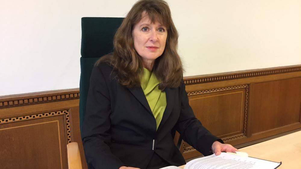 Coroner Jacqueline Lake dressed in green blouse and black jacket, sits at her desk with a book of notes. Behind her is wood panelling with ornate marquetry and a white painted wall above that.