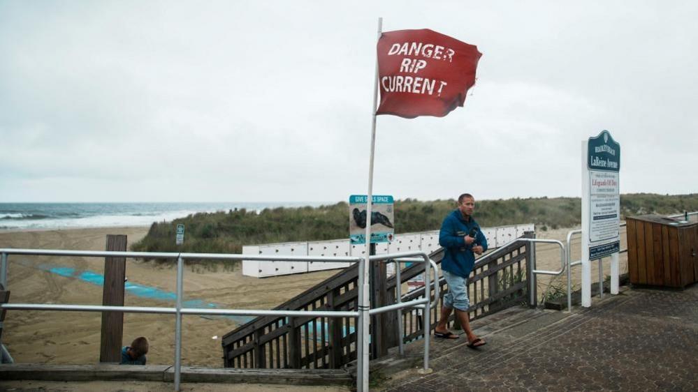 A man in a sweat-suit and flip flops walks away from stairs leading to a beach in Avon-by-the-Sea, New Jersey. Next to him his a red flag reading 'Danger Rip Currents'. The sky ion the background is overcast as Hurricane Erin nears the east coast.