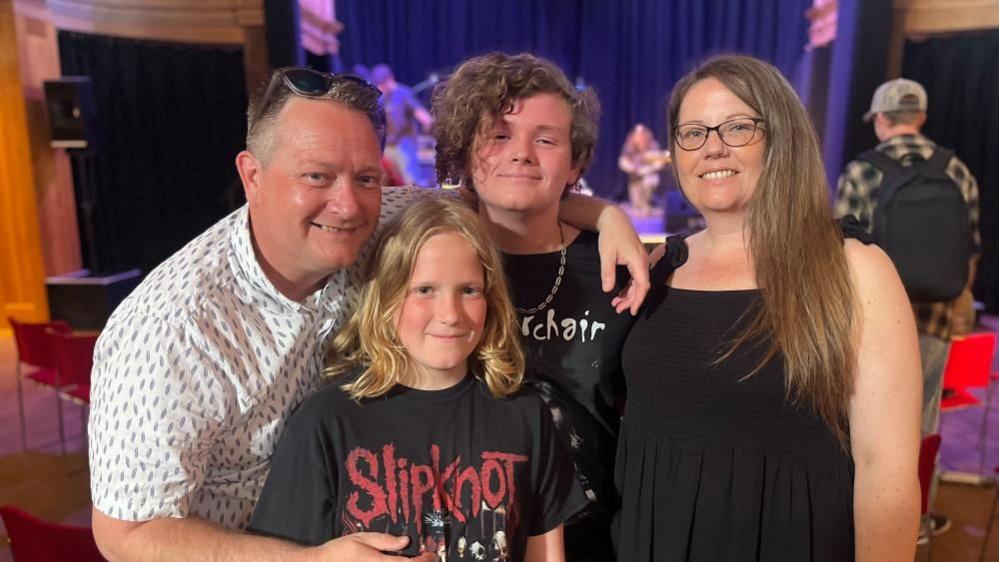 Mark stands to left, with Rupert, then Harry and Holly on the right. Mark is wearing a white patterned shirt, and has sunglasses on his head. Harry is in a black tee shirt bearing the word "Slipknot". He has long blond hair. Behind him is his elder brother Harry, who has medium length brown hair. He is wearing a black shirt with the motif undecipherable. The boys' mum Holly is wearing a black dress. She wears glasses and has long dark blonde hair.