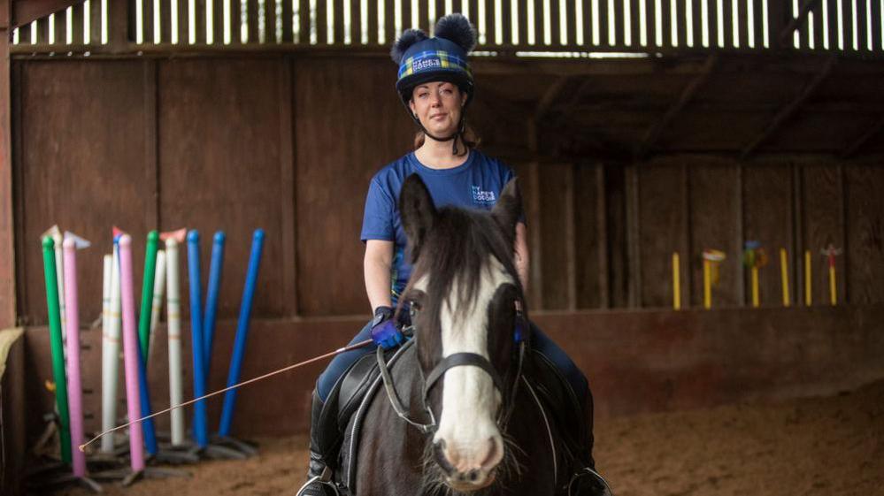 Sheree sits astride a dark brown horse in a riding hall. She wears a blue and yellow tartan My Name's Doddie hat and a blue top, jeans and riding boots as well.