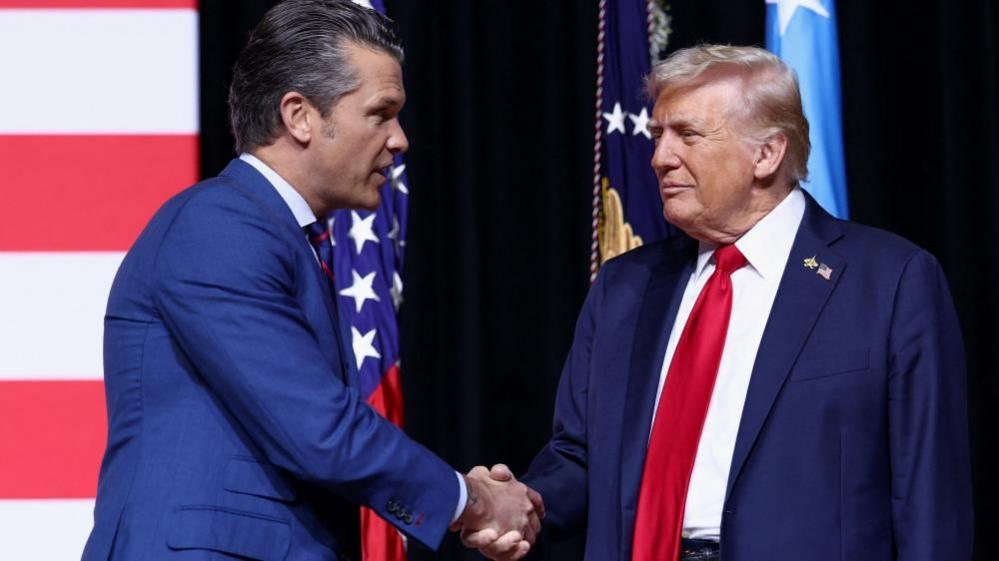 Pete Hegseth shakes Donald Trump's had while standing in front of a US flag