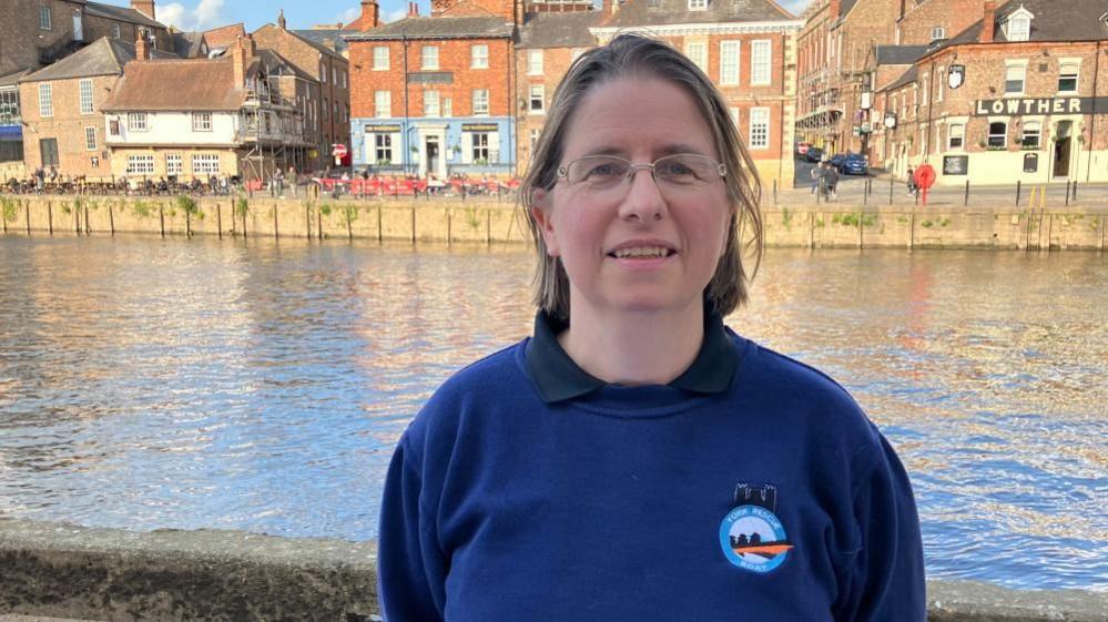 A woman with brown hair wears glasses and a sweatshirt reading "York Rescue Boat". She is standing on Queens Staith in York with the River Ouse in the background.