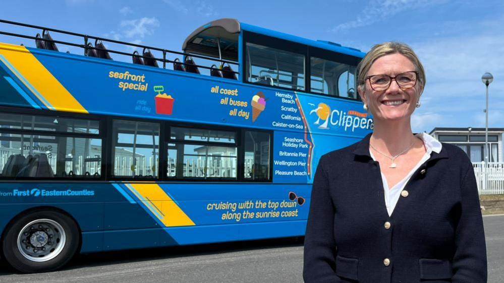 Claire Quinton-Tulloch has long blonde hair tied back, and is wearing glasses. She is wearing a white blouse and a black knitted cardigan over, featuring gold buttons. She is standing next to the Coastal Clipper Cabriolet bus, which is painted in multiple shades of blue, with a yellow diagonal stripe ahead of the rear wheel, sloping towards the back upper deck.