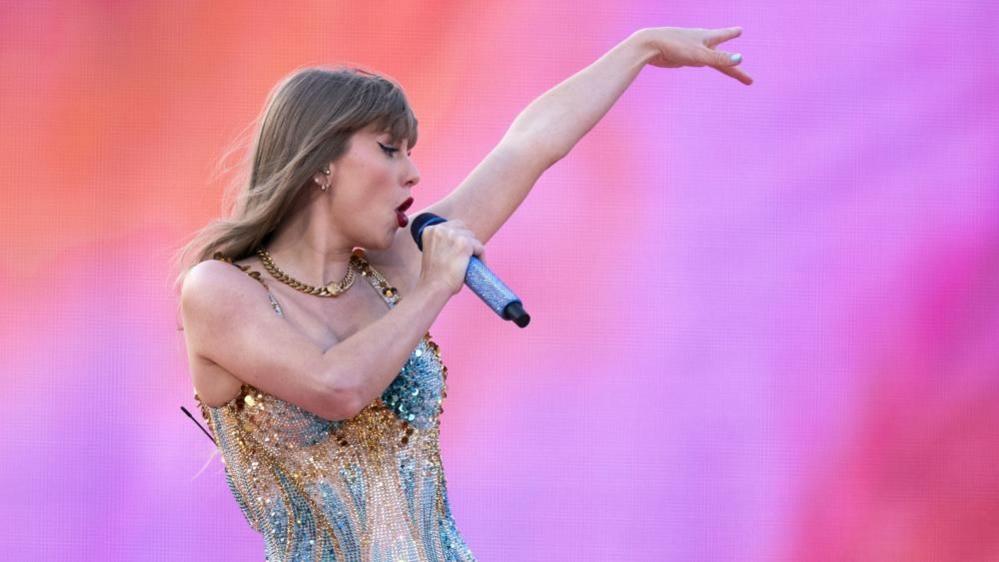 Taylor Swift wearing a sparkly outfit singing on stage in front of a pink and orange backdrop.