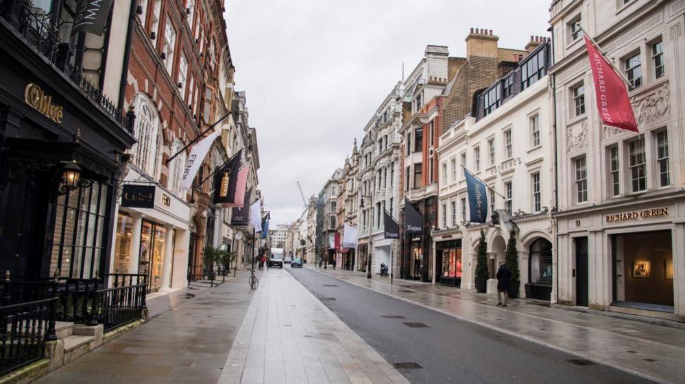 An empty Bond Street, looking down the road on a grey, damp day. Shops line the street on either side, with a wide pavement and a single-lane road through the middle. There are a couple of pedestrians walking along the pavement, and a van and taxi visible in the background.