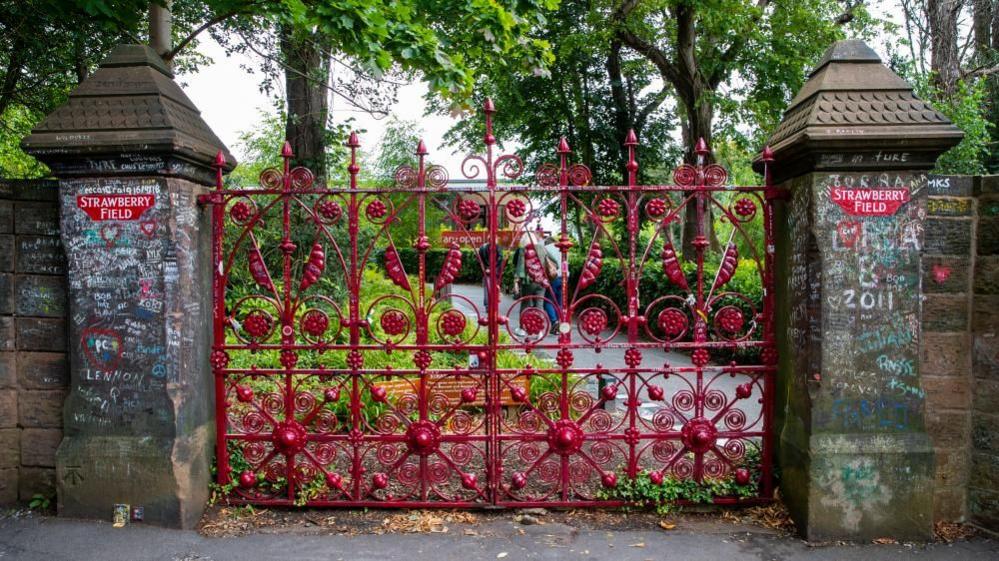 A large red ornate gate is in front of a lush green garden with a path in the middle. Brickwork on either side of the gate is decorated with coloured, hand-drawn messaged and red plaques which say 'Strawberry Field".