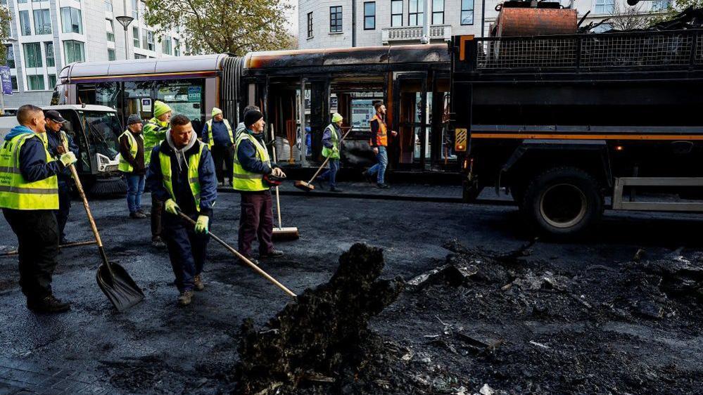 Workers in high viz vests sweep and shovel away the remains of burned vehicles in Dublin city centre on 24 November 2023, the day after the riots. The wreckage of a burnt out Luas tram is in the background.