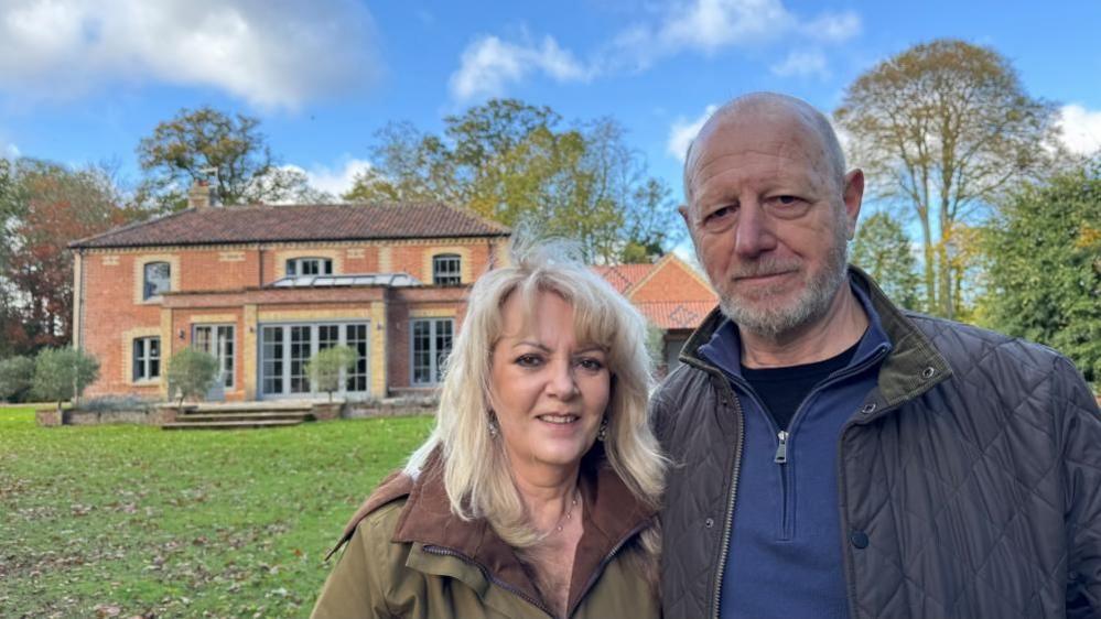 Rachel has long blonde hair and is wearing a wax jacket, with a brown collar. Grenville has close cropped greying hair, a grey beard and is wearing a black tee shirt, with blue zip-fronted sweater and a quilted blue jacket over. They are standing in front of the house where they live, a Victorian building with modern but in-style extensions.
