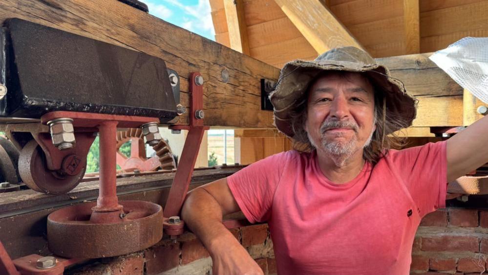 Paul Kemp standing inside the cap of the wind pump, wearing a pink tee shirt and wax hat. He has a greying beard and has one arm resting on the brickwork.