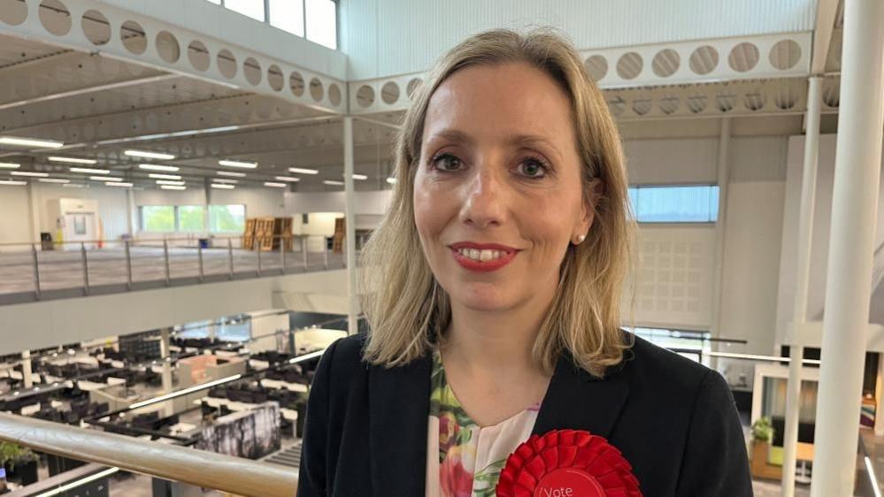 A smiling Sarah Taylor looks directly at the camera, she has shoulder length blonde hair, wearing a floral top and black jacket with a red rosette. She is standing in a building which is white and has a large atrium behind her.