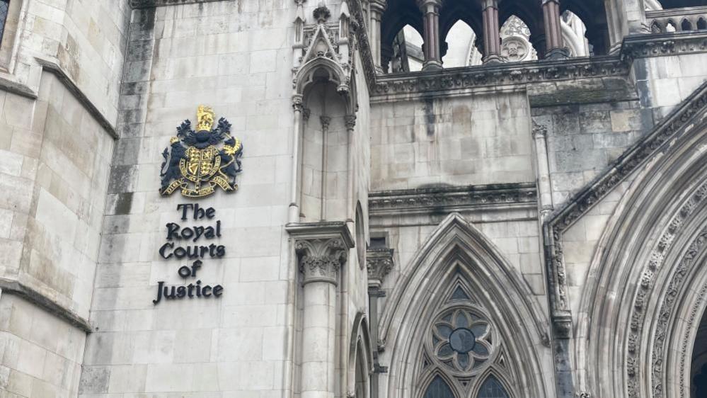 The Royal Courts of Justice: part of a grey stone building. In the top left hand corner are the words "The Royal Courts of Justice", written in black beneath a gold crest.