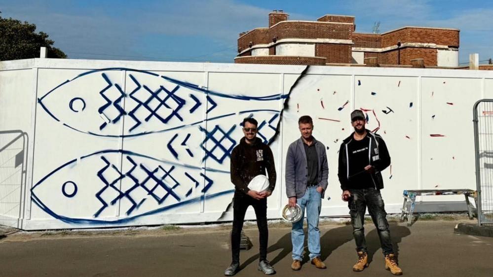 Three men stand by the hoarding outside the Iron Duke pub. The hoarding is painted white with three herring painted in blue depicted.