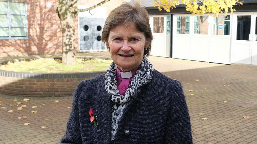 Bishop Olivia Graham stood in courtyard. She has short brown hair and is wearing grey coat over a purple shirt and dog collar.