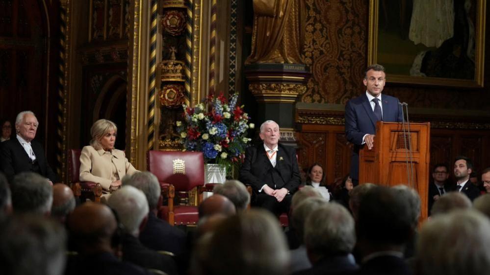 Macron stands at a lectern inside Parliament. Behind him, Speaker Sir Lindsay Hoyle sits with his hands folded in his land, and to his left sits Brigitte Macron.