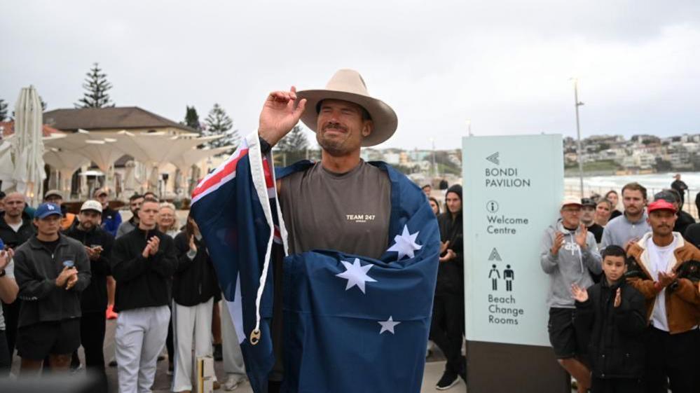 British ultra endurance runner William Goodge, becomes emotional after crossing the finish line at Bondi Beach in Sydney. He is surrounded by supporters and draped in an Australian flag.