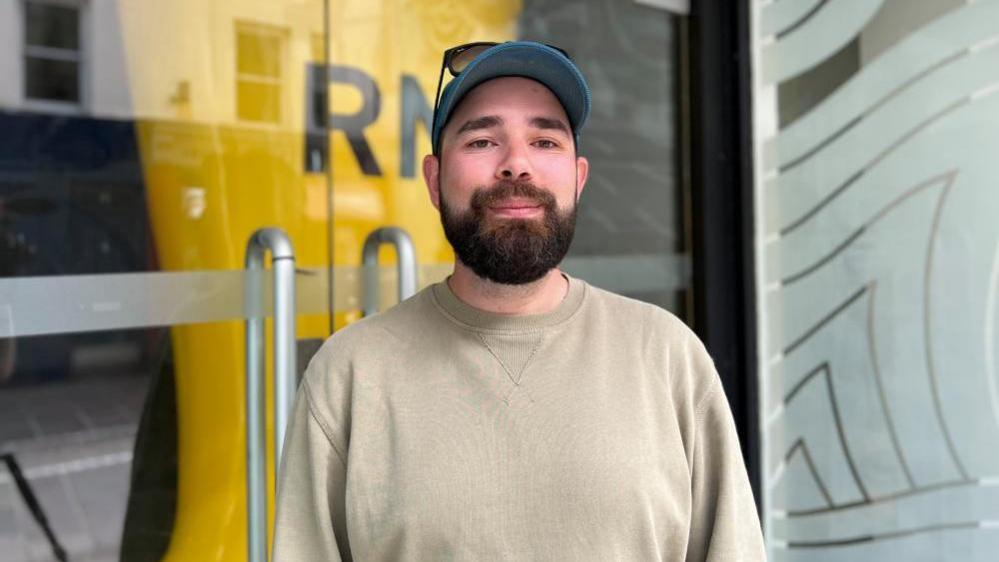 Artist, Theo Jenner stands in front of the shop housing the exhibition of artwork 