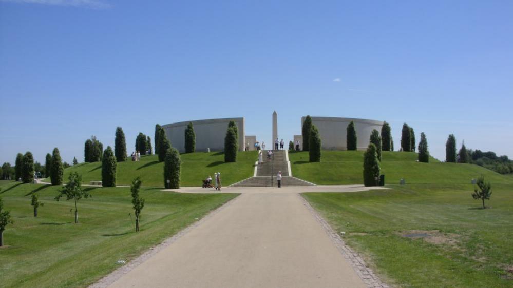 A pathway leading to a war memorial. The memorial is has a stone tower in the centre that comes to a point, which is sounded by circular walls that have an walkway through the middle. A stone path leads to some stone steps that take you up to the memorial. Several trees and larges patches of grass surround the bottom of the memorial.