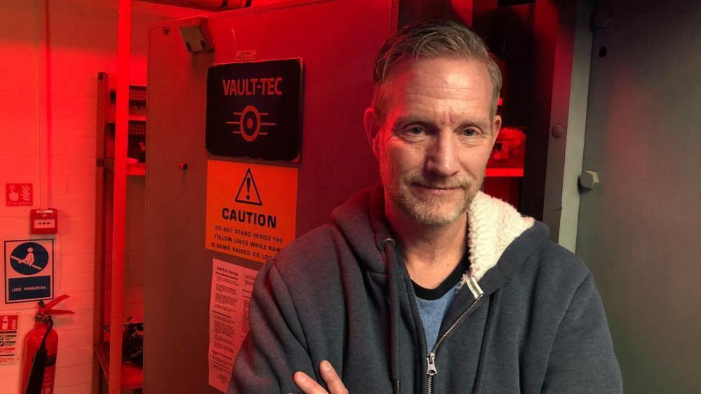 Jon Betts is standing in front of a bank vault door. He is wearing a blue hooded top and has his arms folded. 