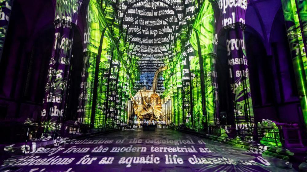 The nave of Salisbury Cathedral lit up for the Sarum Lights. Words are projected against the floor, ceiling and columns with green and purple backlighting.