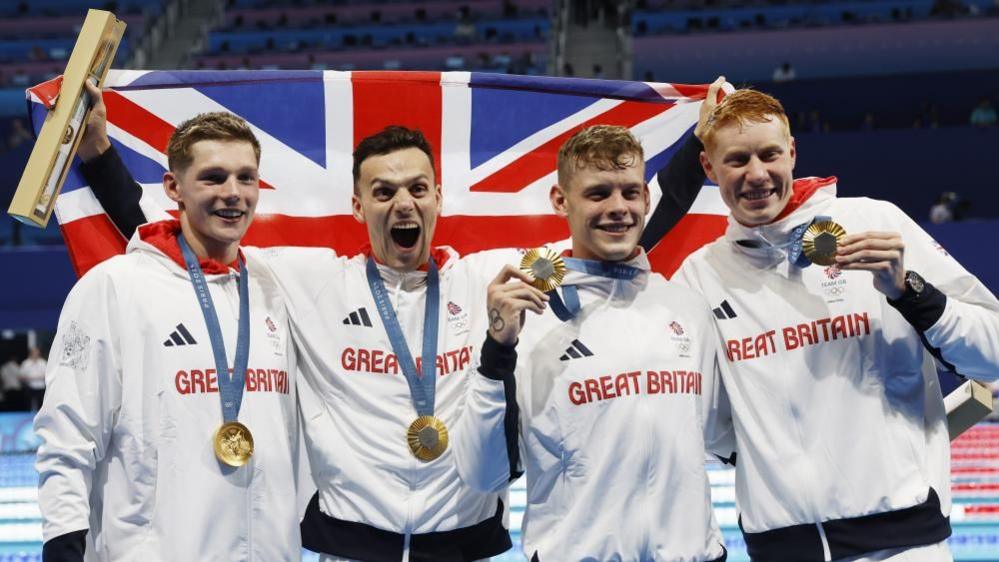 Gold medalist team Britain (L-R) Duncan Scott, James Guy, Matthew Richards, and Tom Dean pose after the Men 4x200m Freestyle Relay final of the Swimming competitions in the Paris 2024 Olympic Games. They are all holding their medals and James is holding up a Union flag and smiling