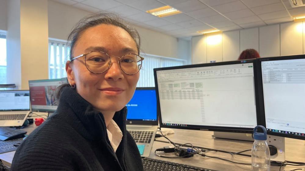 Lizzie Nicolson-Lai is sitting at a desk with computer screens displaying accountancy forms. She is wearing a white shirt under a dark fleece; she is smiling, has her hair tied back and is wearing round-rimmed tortoiseshell glasses and gold hoop ear rings.