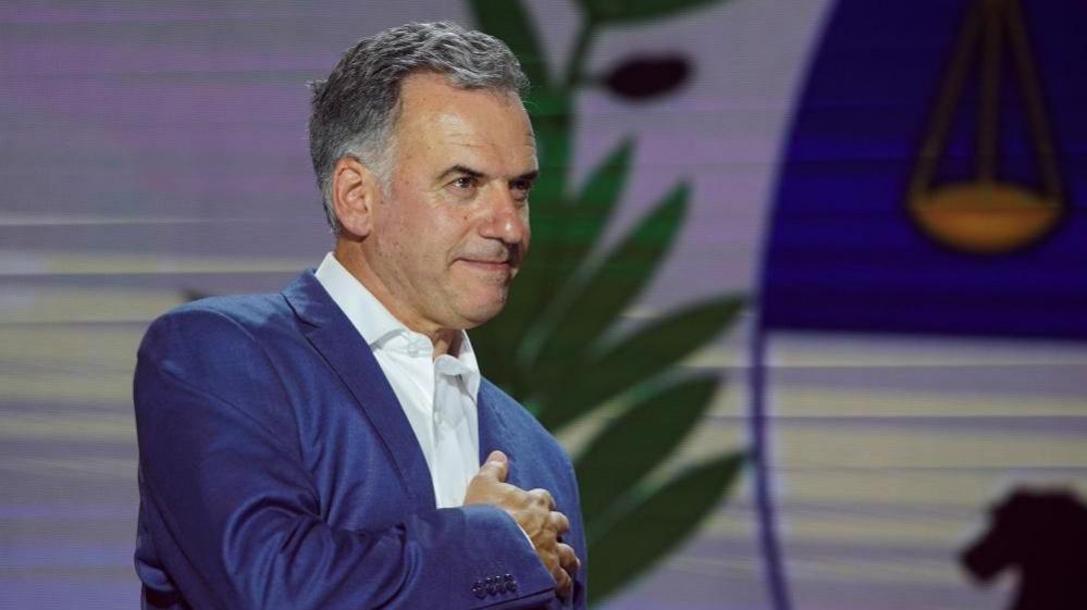 Yamandú Orsi, wearing a blue suit and a white shirt, places his right hand on his heart in a gesture to thank his supporters as early results come in on election night. 